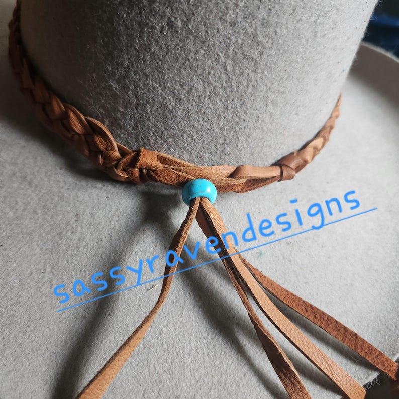 May include: Close-up of a gray hat with a braided brown leather band. A turquoise bead and brown leather tassels hang from the band. The text "sassyravenesigns" is visible in the image.