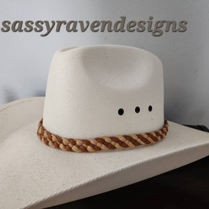 May include: A white cowboy hat with a braided brown and tan band around the crown. The hat has three black dots above the brim. The text "sassyravendesigns" is above the hat.