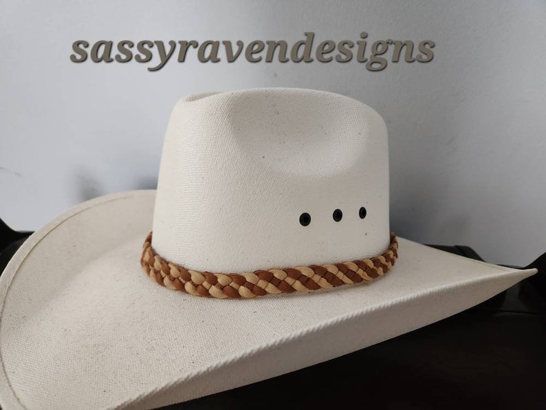 May include: A white cowboy hat with a braided brown and tan band around the crown. The hat has three black dots above the brim. The text "sassyravendesigns" is above the hat.