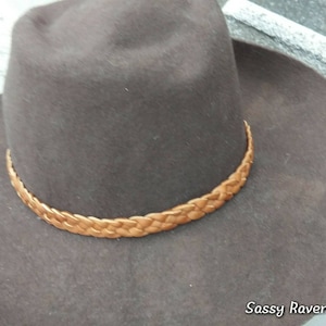 May include: A brown felt hat with a braided leather band. The hat has a wide brim and a classic cowboy hat style. The hat is a neutral color and would be suitable for many outfits.