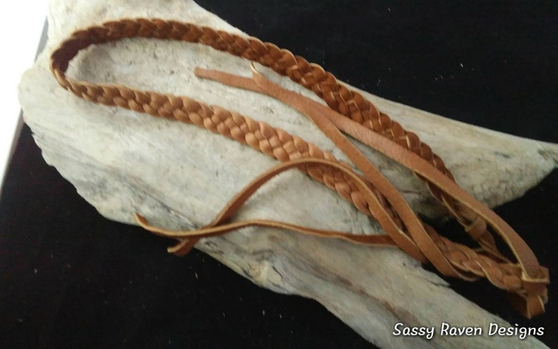May include: A light brown leather headband with a braided design. The headband is lying on a piece of driftwood. The text "Sassy Raven Designs" is in the bottom right corner.