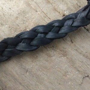 May include: Close-up of a dark blue braided leather strap. The braid is tightly woven, and the leather has a slight sheen. The strap is set against a light brown, textured wooden surface.