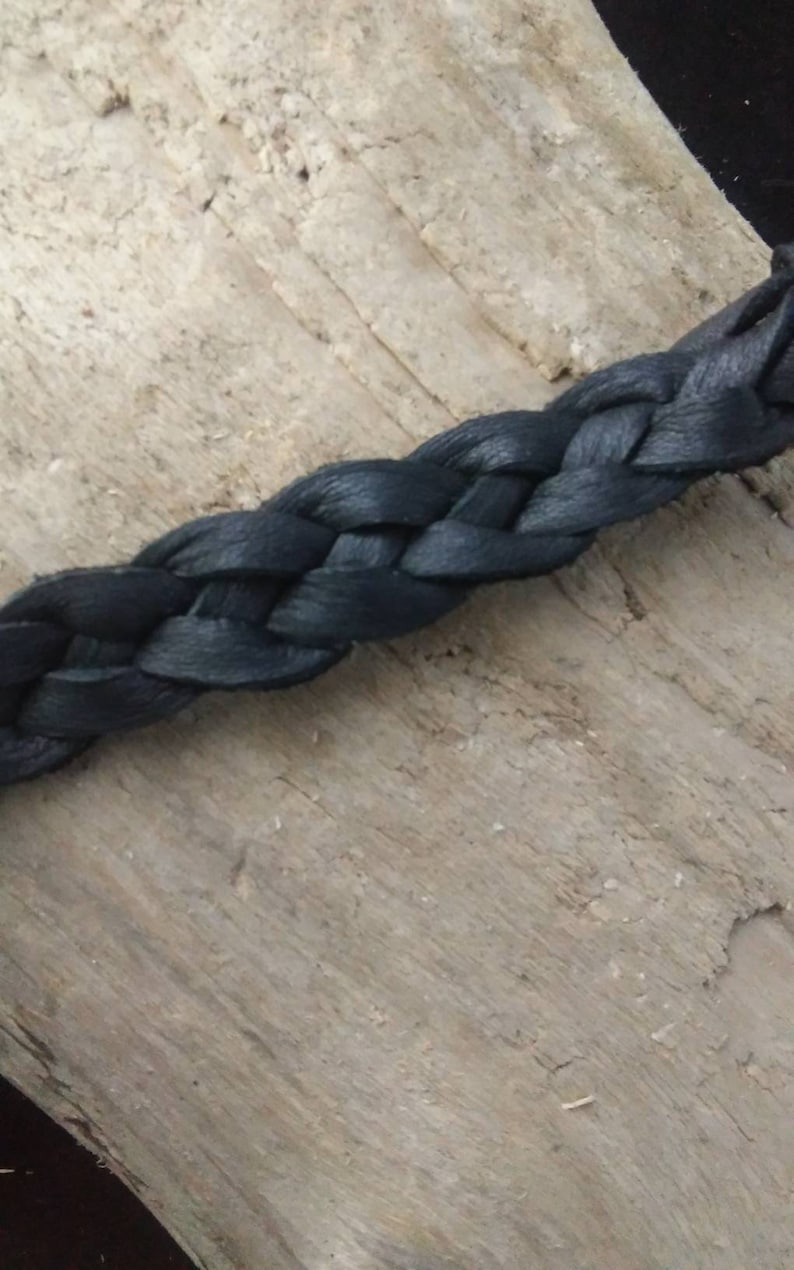 May include: Close-up of a dark blue braided leather strap. The braid is tightly woven, and the leather has a slight sheen. The strap is set against a light brown, textured wooden surface.
