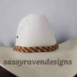 May include: A white cowboy hat with a braided brown and tan band. The hat has a wide brim and a classic western design. The text "sassyravendesigns" is visible on the brim.