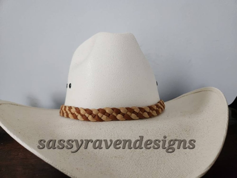 May include: A white cowboy hat with a braided brown and tan band. The hat has a wide brim and a classic western design. The text "sassyravendesigns" is visible on the brim.