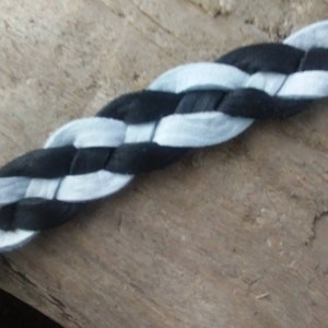 May include: A close-up of a braided cord featuring alternating black and light blue sections. The cord is made of a soft, fabric-like material and is displayed against a natural wood background. The braid is tightly woven.