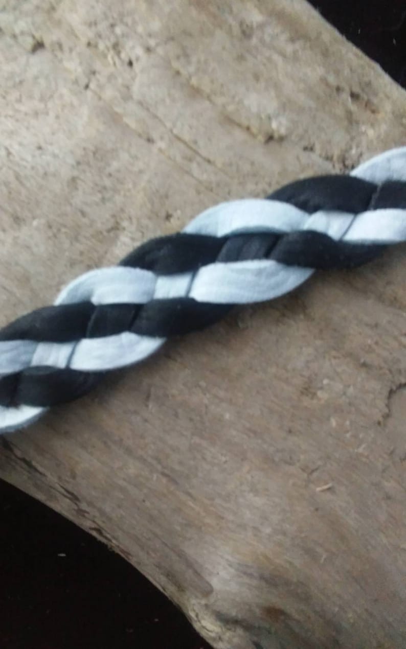 May include: A close-up of a braided cord featuring alternating black and light blue sections. The cord is made of a soft, fabric-like material and is displayed against a natural wood background. The braid is tightly woven.