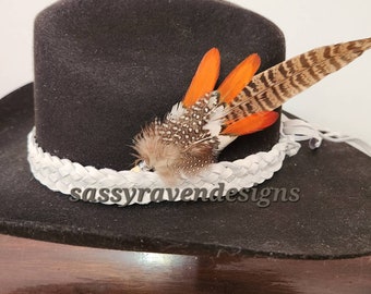May include: A black cowboy hat with a white braided band and a brown and orange feather accent. The hat is sitting on a wooden surface. The text "sass yra vendesigns" is visible on the hat.