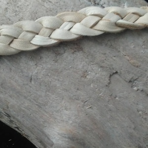 May include: Close-up of a braided leather strap in a natural, light beige color. The strap is made of interwoven strips of leather, creating a textured pattern. The background is a weathered, gray wooden surface.