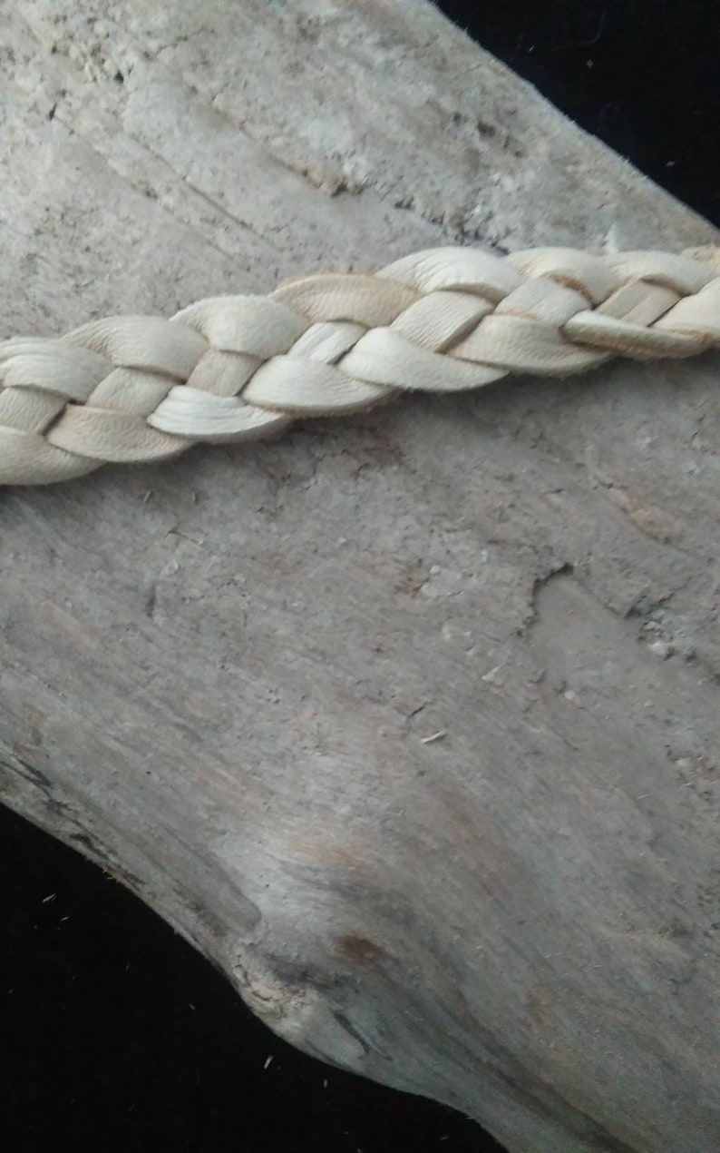 May include: Close-up of a braided leather strap in a natural, light beige color. The strap is made of interwoven strips of leather, creating a textured pattern. The background is a weathered, gray wooden surface.