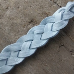 May include: A light blue braided leather strap. The strap is made of woven strips of soft leather. The background is a weathered wooden surface. The strap is approximately 1 inch wide.