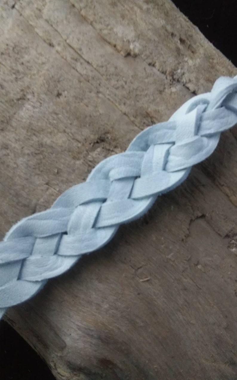 May include: A light blue braided leather strap. The strap is made of woven strips of soft leather. The background is a weathered wooden surface. The strap is approximately 1 inch wide.