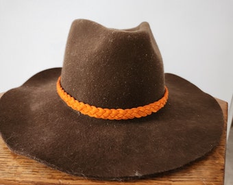 May include: A brown felt cowboy hat with a wide brim and an orange braided band around the crown.