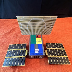 Könnte beinhalten: Ein Modell-Satellit mit Sonnenkollektoren und dem Wort "PROPULSION" auf der Vorderseite. Der Satellit hat einen zentralen Körper mit den Wörtern "FLIGHT CPU", "CAMERA", "BATTERY", "IRIS RADIO" darauf. Die Sonnenkollektoren sind schwarz und gold.