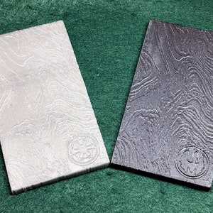 Könnte beinhalten: Zwei rechteckige Holz-Untersetzer mit einer strukturierten Oberfläche. Die Untersetzer sind in verschiedenen Grautönen gehalten. Jeder Untersetzer hat ein kreisförmiges Logo mit einem Sternburst-Design.