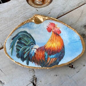 Puede incluir: Un cuenco decorativo de concha con borde dorado. El interior presenta una colorida ilustración de un gallo sobre un fondo azul claro. El gallo tiene plumas rojas, naranjas y azules.