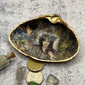 Puede incluir: Una concha decorativa con un borde pintado de oro, que presenta una imagen vintage de una mujer con flores. La concha está rodeada de monedas, cristales y una pequeña botella de vidrio con arena.