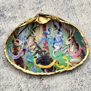 May include: A decorated seashell with a gold-painted edge. The inside displays a colourful illustration of rabbits dancing around a pole with ribbons. The rabbits are in various colours, including brown, black, and white, set against a green background.