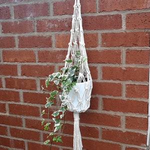 May include: A cream macramé plant hanger with a white ceramic pot containing a green and white plant. The hanger is suspended from a metal ring against a red brick wall.