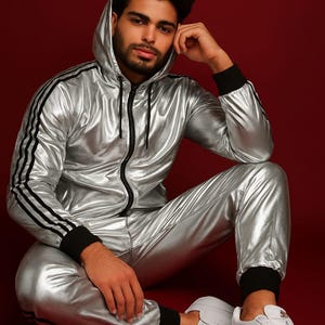 Puede incluir: Chándal plateado con rayas negras en las mangas y las piernas. La chaqueta con capucha tiene cremallera y puños negros. Los pantalones a juego tienen puños negros. Se usan zapatillas blancas. El fondo es rojo liso.