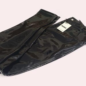 May include: Black, shiny, faux leather trousers folded on a light pink surface. The trousers have a high-waisted design with a belt loop and a tag that reads "RUMO". The material has a glossy appearance.