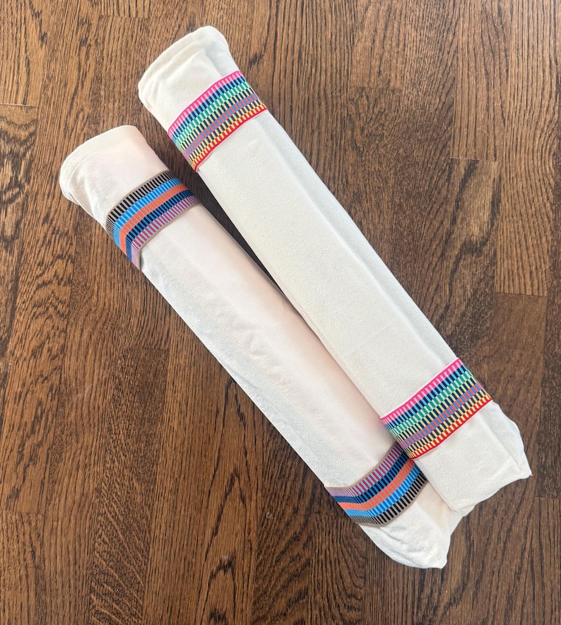 Puede incluir: Dos rollos de tela color blanco hueso asegurados con bandas decorativas. Un rollo presenta una banda con rayas de arco&iacute;ris vibrantes, mientras que el otro est&aacute; sujeto por una banda con rayas horizontales en tonos azul, naranja y negro.
