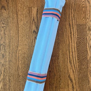 Puede incluir: Un rollo de tela azul claro sujeto por dos bandas el&aacute;sticas. Cada banda presenta un patr&oacute;n repetitivo de finas rayas verticales en colores negro, azul claro, naranja y beige. La tela es lisa y tiene una forma cil&iacute;ndrica bien sujeta.