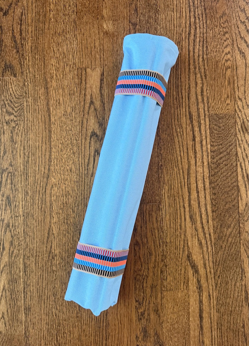 Puede incluir: Un rollo de tela azul claro sujeto por dos bandas el&aacute;sticas. Cada banda presenta un patr&oacute;n repetitivo de finas rayas verticales en colores negro, azul claro, naranja y beige. La tela es lisa y tiene una forma cil&iacute;ndrica bien sujeta.