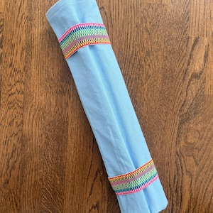 Puede incluir: Un art&iacute;culo de tela azul claro enrollado en un cilindro y asegurado con dos bandas el&aacute;sticas. Cada banda presenta un patr&oacute;n repetitivo de finas rayas verticales en colores vibrantes, incluyendo rojo, amarillo, verde, azul y morado sobre una base blanca.