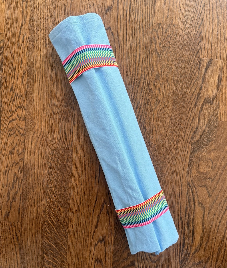 Puede incluir: Un art&iacute;culo de tela azul claro enrollado en un cilindro y asegurado con dos bandas el&aacute;sticas. Cada banda presenta un patr&oacute;n repetitivo de finas rayas verticales en colores vibrantes, incluyendo rojo, amarillo, verde, azul y morado sobre una base blanca.
