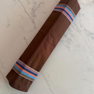 Puede incluir: Un rollo de tela largo y rectangular de color marr&oacute;n, asegurado con dos bandas el&aacute;sticas. Cada banda presenta un patr&oacute;n repetitivo de finas rayas verticales en tonos azul claro, naranja y azul oscuro sobre un fondo beige.