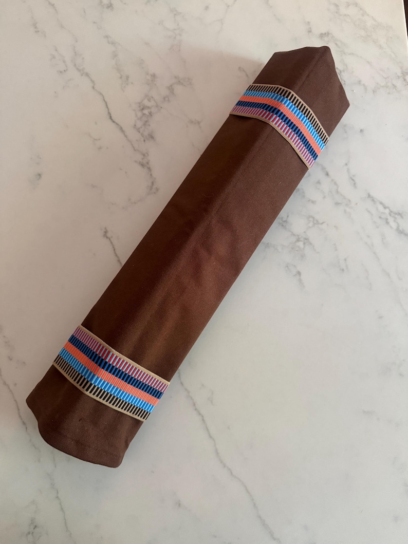 Puede incluir: Un rollo de tela largo y rectangular de color marr&oacute;n, asegurado con dos bandas el&aacute;sticas. Cada banda presenta un patr&oacute;n repetitivo de finas rayas verticales en tonos azul claro, naranja y azul oscuro sobre un fondo beige.