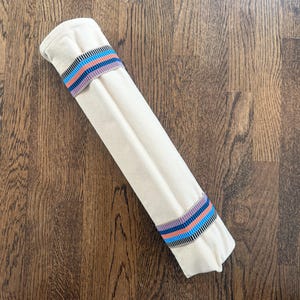 Puede incluir: Un rollo de tela cil&iacute;ndrico de color blanco roto sujeto por dos bandas el&aacute;sticas. Cada banda presenta un patr&oacute;n repetitivo de rayas horizontales en tonos azul brillante, rosa coral y negro, que contrastan con el tejido liso y claro.