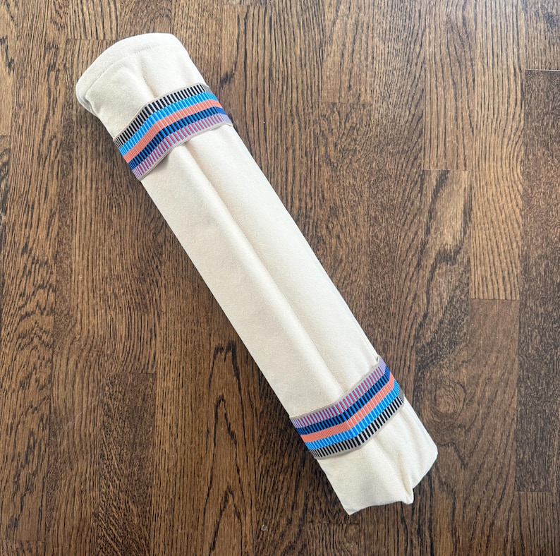 Puede incluir: Un rollo de tela cil&iacute;ndrico de color blanco roto sujeto por dos bandas el&aacute;sticas. Cada banda presenta un patr&oacute;n repetitivo de rayas horizontales en tonos azul brillante, rosa coral y negro, que contrastan con el tejido liso y claro.