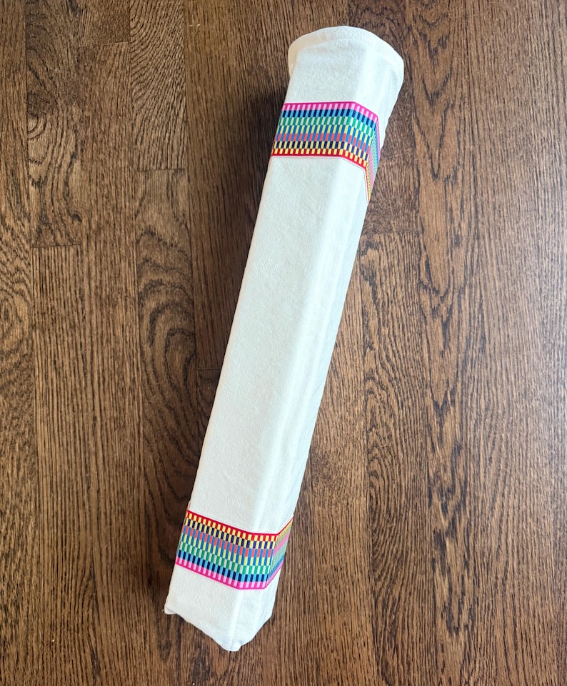 Puede incluir: Un tubo de tela cil&iacute;ndrico de color blanco roto que presenta dos bandas horizontales de patrones tejidos vibrantes y multicolores. Las bandas consisten en peque&ntilde;os cuadrados repetitivos en tonos rojo, naranja, amarillo, verde, azul y morado, que rodean el tejido.