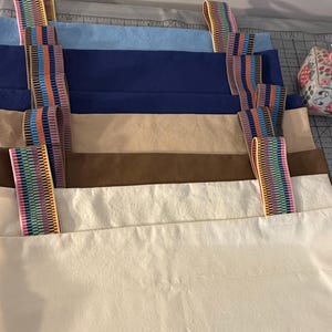 Puede incluir: Una pila de bolsas de tela rectangulares en varios colores s&oacute;lidos, incluyendo azul claro, azul oscuro, beige, marr&oacute;n y blanco roto. Cada bolsa cuenta con dos correas verticales hechas de cinta tejida con patr&oacute;n de arco&iacute;ris.