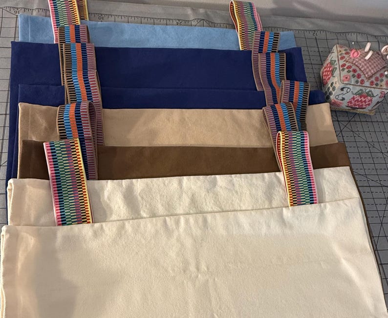 Puede incluir: Una pila de bolsas de tela rectangulares en varios colores s&oacute;lidos, incluyendo azul claro, azul oscuro, beige, marr&oacute;n y blanco roto. Cada bolsa cuenta con dos correas verticales hechas de cinta tejida con patr&oacute;n de arco&iacute;ris.