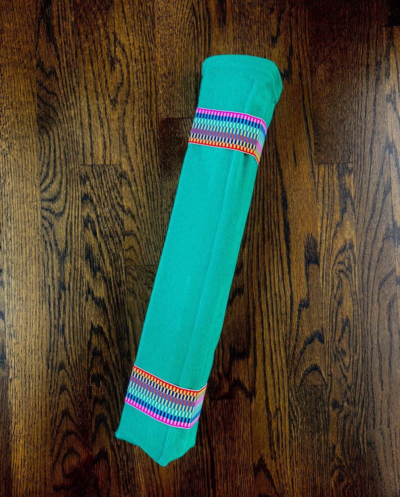 Puede incluir: Una pieza de tela color turquesa s&oacute;lido enrollada y asegurada por dos bandas de cinta tejida. Cada cinta presenta un patr&oacute;n geom&eacute;trico repetitivo en colores vibrantes de arco&iacute;ris, incluyendo rojo, naranja, amarillo, verde, azul y violeta.