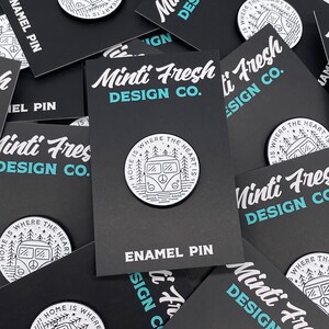 May include: A white enamel pin with a black outline of a vintage camper van with trees and the text "Home is where the heart is" around it. The pin is on a black card with the text "Minti Fresh Design Co." and "Enamel Pin" in white.