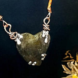 May include: A heart-shaped pendant necklace with a natural, olive-green stone. The stone is accented with silver-colored metal and hangs from a brown leather cord. The necklace has a unique, handcrafted appearance.