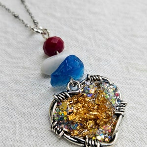 May include: A silver chain necklace with a heart-shaped pendant. The pendant is filled with gold glitter and iridescent glitter. The pendant is surrounded by a silver wire frame. The necklace also features a red bead, a white bead, and a blue bead.
