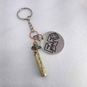 May include: A silver-colored keychain with a round charm featuring a target design and geometric shapes. The keychain includes a chain, copper-colored accents, green beads, and a gold-colored cylindrical charm. The background is a white, textured surface.