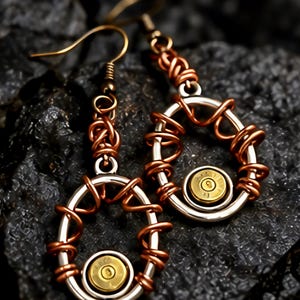 May include: A pair of dangle earrings featuring a central brass-colored bullet casing primer set within a silver-toned oval frame. The frame is wrapped in decorative copper wire, which extends upward to form a coiled link connecting to a hook ear wire.