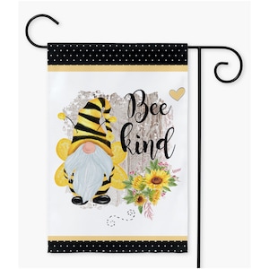 May include: A garden flag with a white background featuring a bee-themed gnome with a yellow and black striped hat and body. The gnome is holding a sunflower and the text "Bee kind" is written in black lettering. The flag has a black and white polka dot border.
