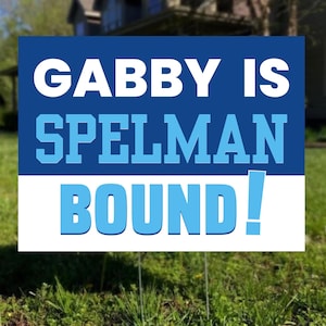 May include: A rectangular yard sign with the text "GABBY IS SPELMAN BOUND!" in white and light blue lettering on a dark blue and white background. The sign is placed in a grassy yard.