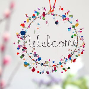 Peut inclure: Une couronne de perles colorées avec le mot "Bienvenue" écrit en fil de fer. La couronne est décorée de perles, de boutons et d'autres petites babioles de différentes couleurs, notamment le rouge, le bleu, le vert, le jaune et le violet.