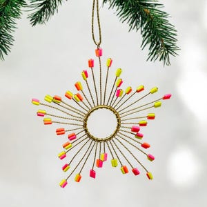Könnte beinhalten: Ein sternförmiger Weihnachtsschmuck mit einem goldfarbenen Drahtrahmen und farbenfrohen Akzenten. Der Schmuck hat einen zentralen goldenen Ring mit radialen Drähten, die jeweils mit rosa, gelben und orangefarbenen Perlen versehen sind. Zum Aufhängen ist eine goldene Schnur befestigt.