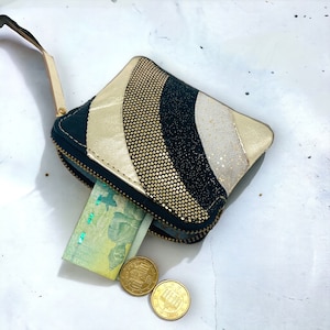 Può includere: Un piccolo portafoglio con cerniera e cinturino da polso dorato. Il portafoglio presenta un design a strisce curve in oro, nero e bianco. Una banconota e due monete sono visibili all'interno e accanto al portafoglio.