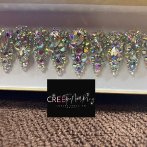 May include: A set of ten iridescent press-on nails with a crystal design. The nails are in a white box with the text "CREE The Nail Plug LUXURY PRESS ON NAILS".