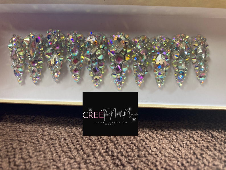 May include: A set of ten iridescent press-on nails with a crystal design. The nails are in a white box with the text "CREE The Nail Plug LUXURY PRESS ON NAILS".